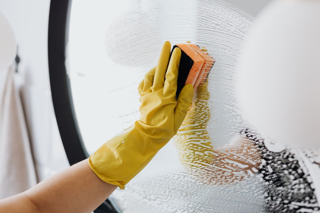 Un individu anonyme portant des gants en caoutchouc jaunes nettoie un miroir de salle de bain avec une simple éponge