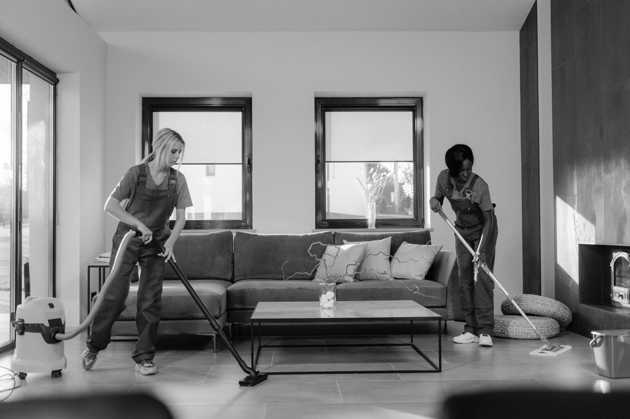 about-01 Two women in work clothes cleaning a modern living room using a vacuum and mop.